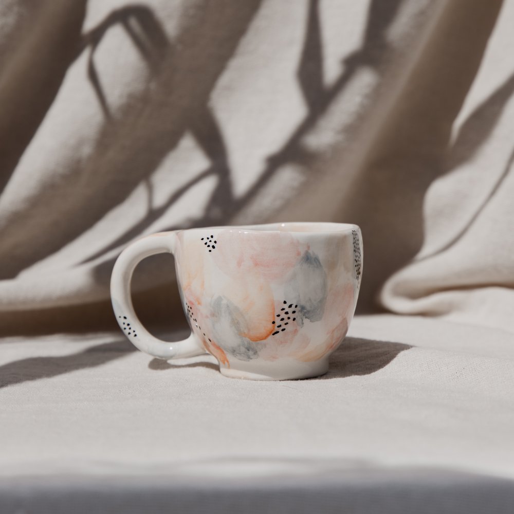 A hand-painted ceramic mug with soft peach and grey brushstrokes on sunlit linen