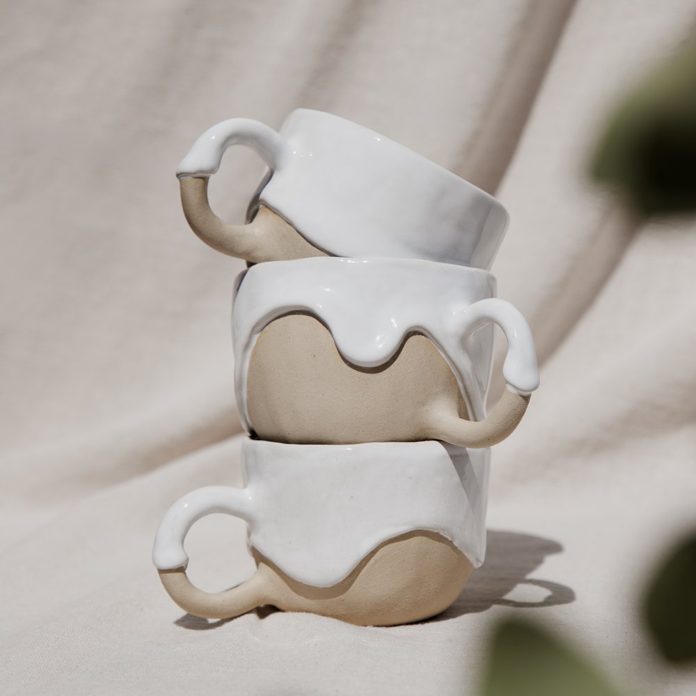 A stack of three organic ceramic mugs with curved handles and dripping white glaze