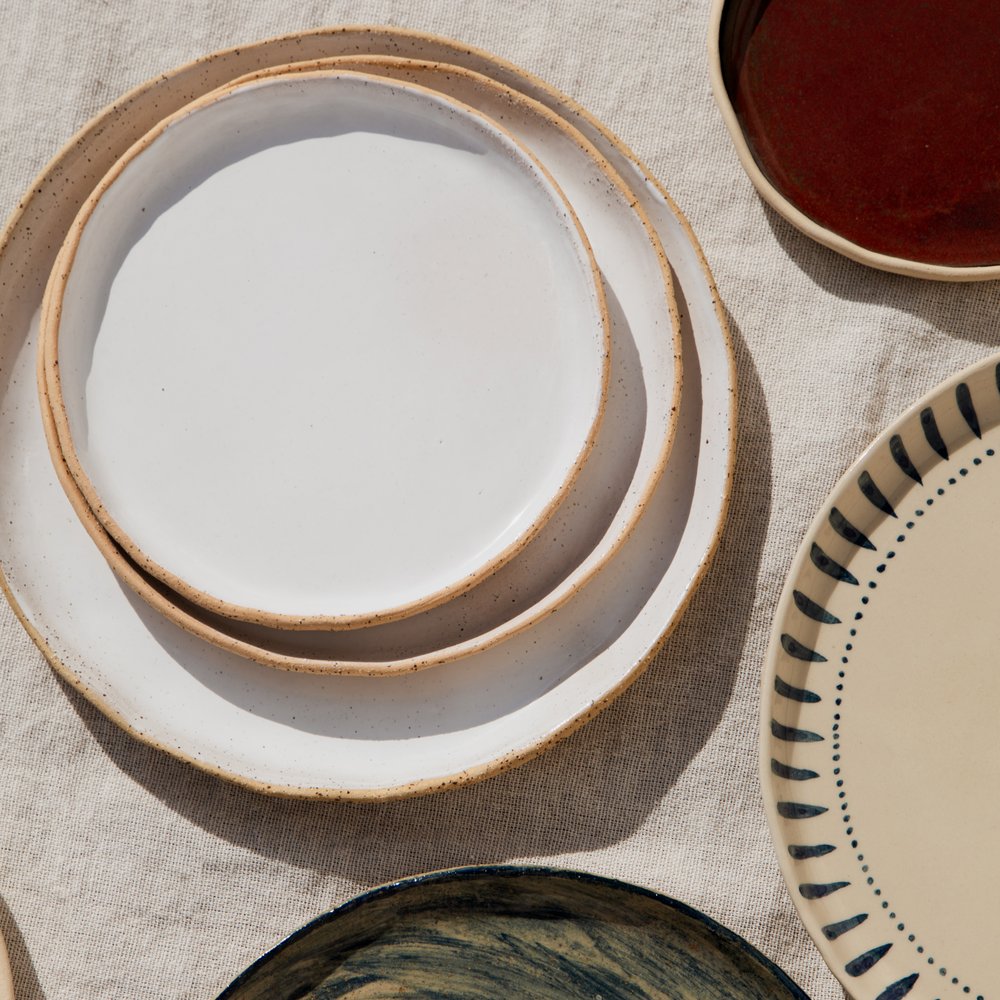 A flat-lay of nested speckled stoneware plates with a deep-red glazed plate and a blue-striped plate alongside