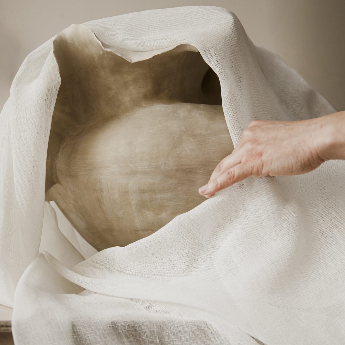 A hand lifting a linen cloth from a freshly formed ceramic vessel left to dry slowly