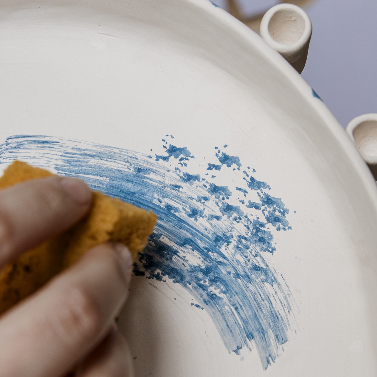 A hand sweeping cobalt pigment across the surface of a hand-built ceramic dish with a sponge